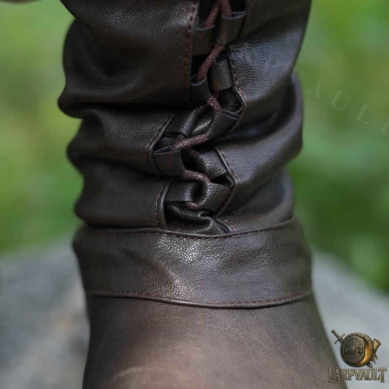 May include: Close-up of a brown leather boot cuff with a laced closure. The leather is textured and has a slightly worn appearance. The cuff is gathered and has a band at the bottom. The laces are a darker brown.