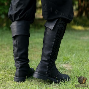 Puede incluir: Botas negras hasta la rodilla con dise&ntilde;o de cordones y pu&ntilde;o doblado. Las botas son de material texturizado y tienen suela plana. Se usan con pantalones negros y sobre un fondo de hierba verde.