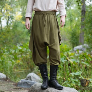 May include: Olive green harem pants with a drawstring waist and a draped side panel. The pants are paired with black lace-up boots. The person is standing on a large rock in a natural outdoor setting.