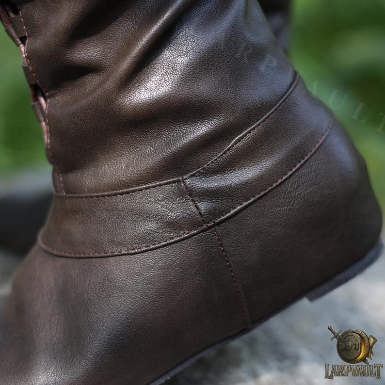 May include: Close-up of a brown leather boot. The boot features a flat sole and a strap detail around the ankle. The leather has a textured appearance, and the stitching is visible. The boot appears to be new.