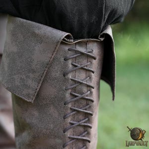 Puede incluir: Primer plano de una bota de cuero marr&oacute;n con un acabado texturizado y un pu&ntilde;o doblado. La bota presenta un cierre de cordones que recorre el lateral. La parte superior de la bota est&aacute; parcialmente oscurecida por tela oscura. La bota est&aacute; dise&ntilde;ada para recreaciones hist&oacute;ricas o cosplay.