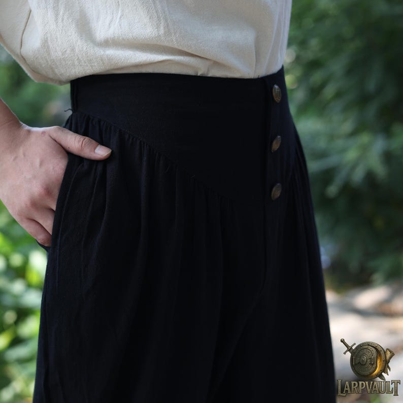 May include: Black high-waisted pants with three decorative buttons and a pocket. The pants are made of a soft, flowing fabric and are paired with a cream-colored top. The pants have a vintage or historical style.