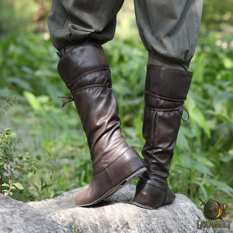 May include: Brown leather knee-high boots with a flat sole. The boots have a folded-over cuff and are laced with a thin brown cord. The boots are worn with olive green pants. The boots are designed for historical reenactment or cosplay.