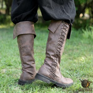 Puede incluir: Botas marrones hasta la rodilla con un dise&ntilde;o de cordones en el lateral. Las botas tienen un pu&ntilde;o doblado en la parte superior y una suela gruesa y negra. Las botas se llevan con pantalones negros.