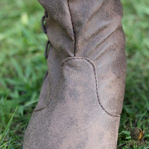 Puede incluir: Primer plano de una bota de cuero marr&oacute;n con un acabado envejecido. La bota presenta un dise&ntilde;o con cordones y detalles de costura visibles. La bota se encuentra sobre un fondo de hierba verde, lo que sugiere un entorno al aire libre.