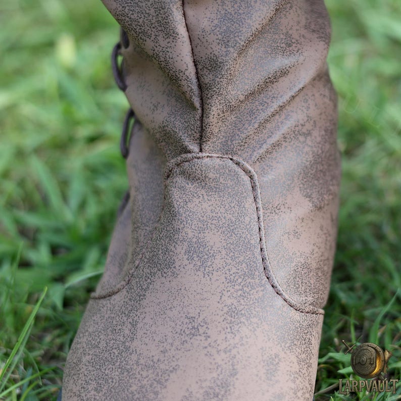 Puede incluir: Primer plano de una bota de cuero marr&oacute;n con un acabado envejecido. La bota presenta un dise&ntilde;o con cordones y detalles de costura visibles. La bota se encuentra sobre un fondo de hierba verde, lo que sugiere un entorno al aire libre.