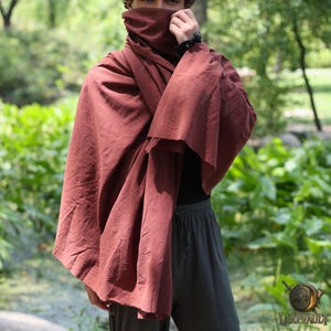 Może przedstawiać: Rdzawo-czerwony płaszcz z wysokim kołnierzem, narzucony na ramiona. Płaszcz wykonany jest z teksturowanej tkaniny i ma płynny krój. Osoba ma na sobie ciemne spodnie i czarną koszulę. Tło to rozmyta sceneria na zewnątrz.