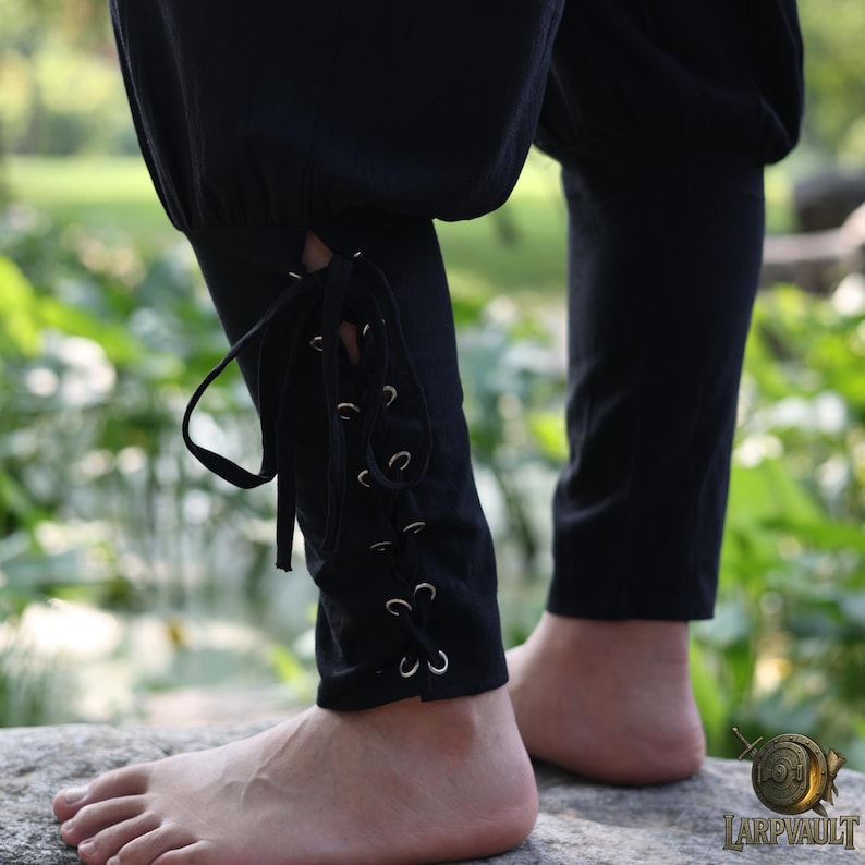 May include: Black pants with lace-up detailing on the lower leg. The pants have a loose fit at the top and taper down to the ankle. The laces are secured with metal eyelets. The person is standing on a rock in a natural setting.