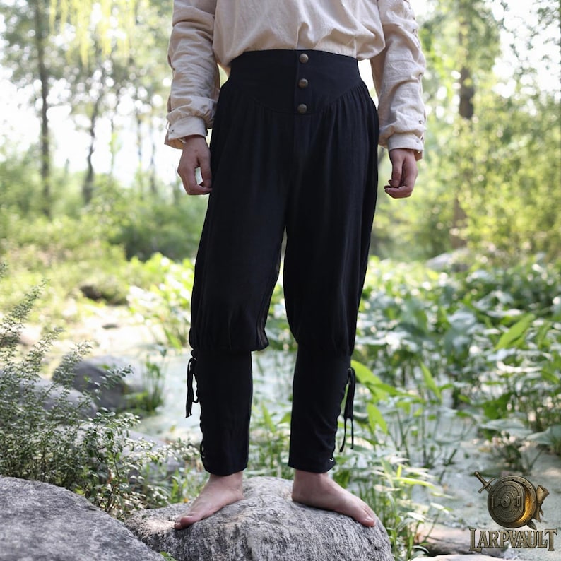May include: Black medieval pants with a high waist and button closure. The pants have a loose fit through the thighs and taper to the ankles, with tie closures at the cuffs. The pants are made of a lightweight fabric.