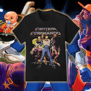 May include: Black t-shirt featuring a colorful graphic of a superhero team with the text "CAPTAIN COMMANDO" in a retro font. The design includes various characters in action poses, with a baby character in the background. The shirt is a classic crew neck.