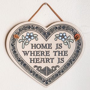 May include: A heart-shaped ceramic wall plaque with a cream-colored base and a dark blue decorative border. The text "HOME IS WHERE THE HEART IS" is printed in the center in dark blue, accompanied by light blue floral accents and a brown hanging cord.