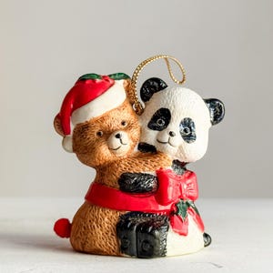 May include: A ceramic Christmas ornament featuring a brown teddy bear wearing a Santa hat and hugging a panda bear. The panda has black and white markings and a red bow. The ornament has a gold string for hanging.