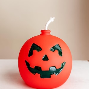 May include: A round, orange Halloween candle shaped like a jack-o'-lantern. The candle has a carved face with black features and a white wick. The candle is a festive decoration for the Halloween season.