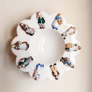 May include: White ceramic serving bowl with a scalloped edge, adorned with small, colorful figurines around the rim. The figurines depict people in various outfits. The bowl has a central, circular indentation.
