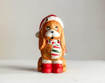 May include: A ceramic dog figurine wearing a Santa hat, scarf, and boots. The dog is brown with a white face and a red and white hat. The scarf has red and green stripes. The boots are red.