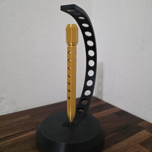 May include: A black and gold headphone stand. The base is a solid black circle, supporting a curved black arm with circular cutouts. A gold-colored vertical post with diamond-shaped cutouts holds the headphones.
