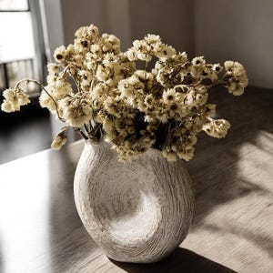 May include: A white textured vase with a unique, organic shape, filled with dried, cream-colored flowers. The vase sits on a wooden surface, creating a neutral and natural aesthetic. The flowers add a touch of rustic charm to the decor.