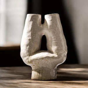 May include: A textured, off-white ceramic vase with a unique, abstract design. The vase features a central opening and two vertical, rounded forms that meet at the top. The base is a solid, rounded shape. The vase is on a wooden surface.