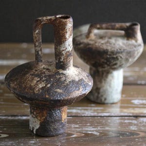 May include: Two handcrafted ceramic vases with a rustic, aged appearance. One vase features a handle, while the other has a rounded top. The vases are primarily brown with white and tan accents, showcasing a textured surface. They are placed on a wooden surface.