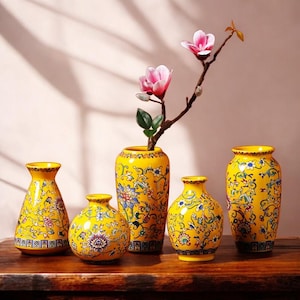 Könnte beinhalten: Fünf gelbe Keramikvasen mit floralen Mustern. Die Vasen haben unterschiedliche Formen und Größen und sind mit blauen, grünen und roten Designs verziert. Ein Zweig mit rosa Blüten befindet sich in einer Vase und fügt dem Arrangement ein natürliches Element hinzu.