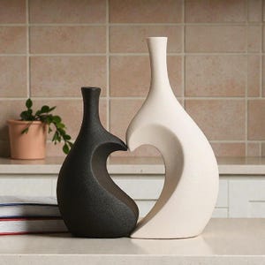 May include: Two ceramic vases, one black and one off-white, stand side-by-side. The black vase has a curved cutout, while the white vase has a flowing, open design. The vases are on a white surface, with a plant and books in the background.