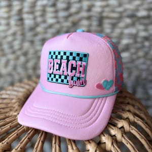 Pode incluir: Boné rosa estilo trucker com aba curva e parte traseira em rede. A frente apresenta a palavra "BEACH" em rosa e preto, com um fundo xadrez. Um pequeno patch de coração e a palavra "babe" também estão presentes. O boné tem uma borda azul claro.