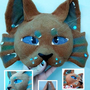 May include: A brown felt cat mask with blue eyes, black nose, and teal fin-like side panels. The mask features small blue gem accents and long whiskers. The text "AQUATIC CAT" appears at the top, and "REVALYWINGS" is at the bottom.