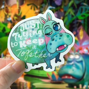 May include: A green and blue watercolor sticker of a cartoon hippopotamus with the text "Just trying to keep it together".