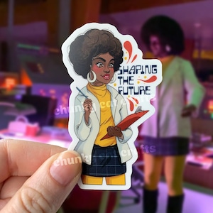 May include: A sticker featuring a cartoon illustration of a woman with an afro wearing a white lab coat and yellow turtleneck. She is holding a clipboard and a pen. The text "SHAPING THE FUTURE" is in red and white with a red and orange swirl design.