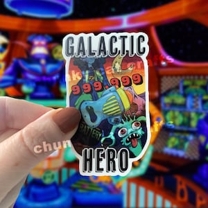May include: A sticker with a colorful illustration of a cartoon character holding a tool with the words "Galactic Hero" and "999.999" printed on it.