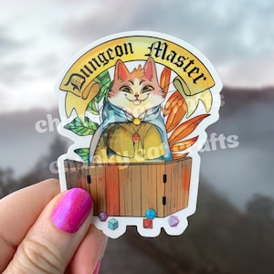 May include: A sticker featuring a cartoon cat dressed as a Dungeon Master. The cat is sitting behind a wooden table with dice scattered around it. The text "Dungeon Master" is written in a scroll-like banner above the cat.