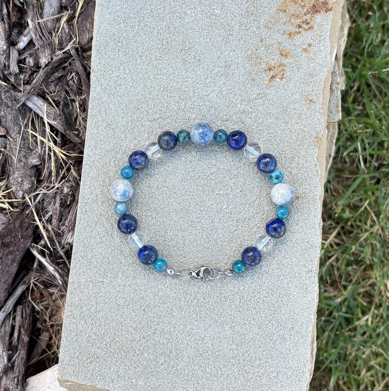 May include: A beaded bracelet featuring a variety of blue and clear beads. The bracelet includes dark blue, light blue, and clear beads, with a silver clasp. The bracelet is displayed on a gray stone surface.