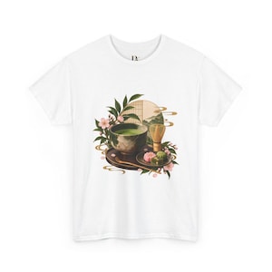 以下が含まれることがあります： 白いTシャツに、緑色の抹茶が入った茶碗、竹製の茶筅、そして暗い色の盆に乗った色とりどりの和菓子が描かれたグラフィックプリント。イラストにはピンクの桜の花、緑の葉、円形の背景要素が含まれています。
