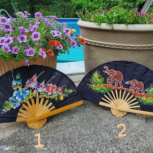 May include: Two black hand fans with wooden handles. One fan features a floral and bird design, the other depicts elephants. The fans are displayed outdoors near potted flowers and a swimming pool.
