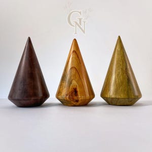 Puede incluir: Tres objetos de madera en forma de cono en tonos marrón, beige y verde. Cada uno tiene una base plana y facetada y una parte superior puntiaguda. Los objetos se muestran sobre una superficie blanca, mostrando la veta y las texturas naturales de la madera.