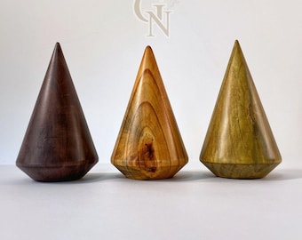 Puede incluir: Tres objetos de madera en forma de cono en tonos marrón, beige y verde. Cada uno tiene una base plana y facetada y una parte superior puntiaguda. Los objetos se muestran sobre una superficie blanca, mostrando la veta y las texturas naturales de la madera.