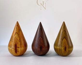 Puede incluir: Tres esculturas de madera en forma de lágrima en tonos marrones y amarillos. Las esculturas tienen un acabado liso y pulido y una parte superior puntiaguda. La veta de la madera es visible, agregando textura. Se exhiben sobre un fondo blanco.