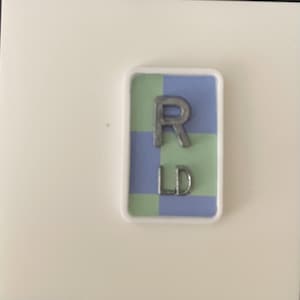 May include: A rectangular sign with a white border, featuring a blue and green checkered background. The letters "R" and "LD" are in silver, metallic lettering. The sign is set against a white background.