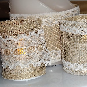 May include: Two cylindrical candle holders made of tan hessian fabric wrapped with white lace trim. The lace features a repeating oval pattern along the top and bottom edges. A warm, glowing light is visible through the mesh of the hessian.