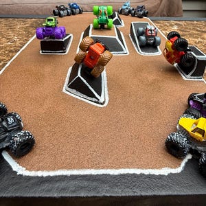 Puede incluir: Una escena de rally de monster trucks en miniatura con varios camiones de juguete coloridos sobre una superficie marrón y negra. Los camiones están en diferentes posiciones, algunos en rampas, otros en posición vertical y otros volcados. La escena está configurada para parecerse a una pista de carreras.
