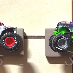 Può includere: Due monster truck giocattolo, uno bianco con dettagli rossi e blu, l'altro verde e nero. Entrambi hanno grandi pneumatici neri con cerchi rossi e sono montati su supporti grigi con ganci.