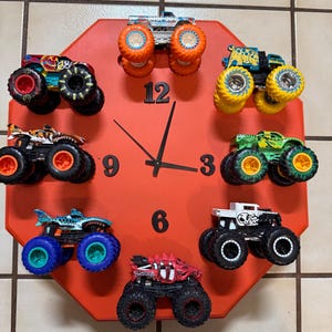 May include: A red octagonal clock with black hands and numbers. Monster truck toys are arranged around the clock face, with one at each hour. The trucks are various colors, including blue, green, and orange.