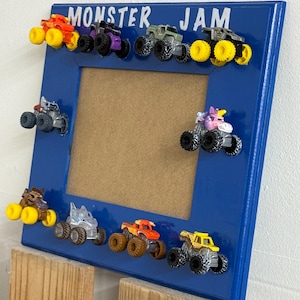 May include: A blue rectangular picture frame featuring the text MONSTER JAM at the top. The frame is decorated with ten miniature monster truck toys in various colours including orange, purple, green, grey, and yellow, attached to the border.