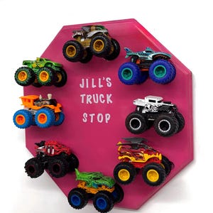 May include: A pink octagonal wall display featuring the white text JILL'S TRUCK STOP. Seven miniature monster trucks in various colours including green, orange, red, yellow, and white are attached to the board in a circular arrangement.