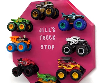 May include: A pink octagonal wall display featuring the white text JILL'S TRUCK STOP. Seven miniature monster trucks in various colors including green, orange, red, yellow, and white are attached to the board in a circular arrangement.