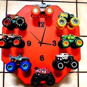 May include: An octagonal red wall clock featuring black numbers 3, 6, 9, and 12, with black clock hands. Seven miniature monster trucks with various colourful designs and oversized wheels are attached to the clock face as hour markers.