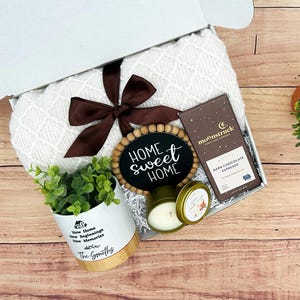 May include: A gift box with a white blanket, a dark chocolate espresso bar, a small potted plant, a candle, and a wooden sign that says 'HOME sweet HOME'. The sign is surrounded by wooden beads. The plant pot is white with 'New Home, New Beginnings, New Memories, The Smiths' printed on it. The box is on a wooden surface.