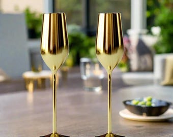 May include: Two gold-colored champagne flutes with a sleek, modern design. The flutes have a tall, slender stem and a wide, rounded bowl. The glasses are made of a shiny, reflective material, and are sitting on a light-colored table.