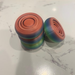 Op de afbeelding: Een stapel van vier kleurrijke, ronde objecten met concentrische cirkels bovenop. De bovenkant van elk object is rood-oranje, overgaand in blauw en groen aan de zijkanten. Eén object is gekanteld en toont het interieurontwerp.
