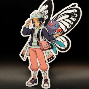 May include: A sticker featuring an anime-style character with butterfly wings, holding a purple creature and a red and white ball. The character wears a hat, jacket, and pink pants. The background is black.